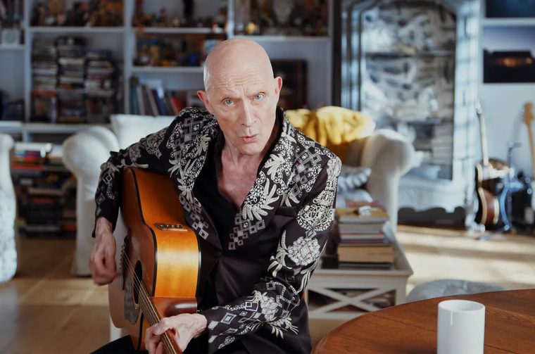 A bald man in a distinctive black shirt with white tropical or pineapple patterns, holding an acoustic guitar in what appears to be a modern, well-appointed living space with bookshelves and contemporary furnishings.