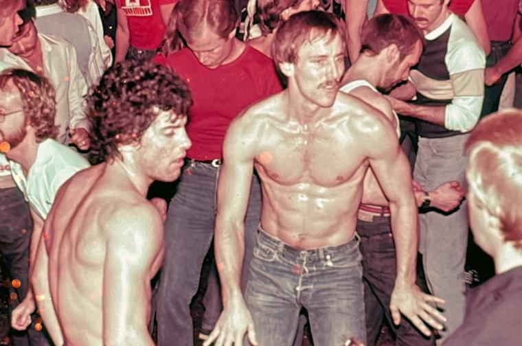Two shirtless white men wearing jeans stand in the middle of a gay nightclub in the 70s, caught in the middle of a dance.