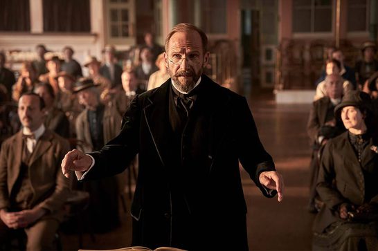 Man in black formal coat and glasses gesturing while stood in front of a seated audience in a church. He is conducting a choir.