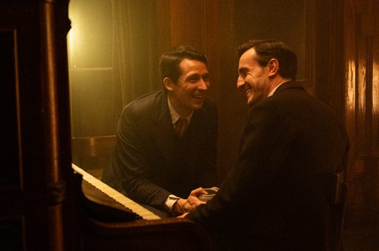 We see two men in formal 1930s or 1940s-era attire sharing an intimate, warm moment at a piano. The scene has a rich, golden-toned lighting that creates an atmosphere of elegance and closeness. One man is seated at the piano while the other leans in close, both smiling genuinely at each other.