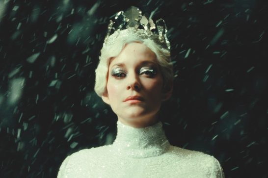 A woman in her forties (Marion Cotillard) with wavy short blinde hair stares into the camera with a serious expression on her face. She has ice blue eye shadow and is wearing a crown made of shards of glass and a white sequinned turtleneck. We see flakes of snow gusting around her.