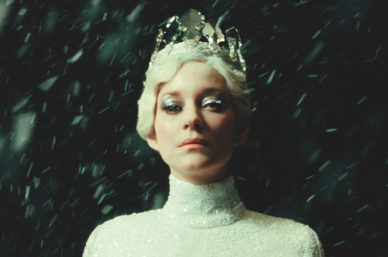 A woman in her forties (Marion Cotillard) with wavy short blinde hair stares into the camera with a serious expression on her face. She has ice blue eye shadow and is wearing a crown made of shards of glass and a white sequinned turtleneck. We see flakes of snow gusting around her.