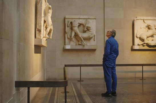An older man in blue overalls stands in a museum, staring up at some marble scupltures that are hanging on the walls.