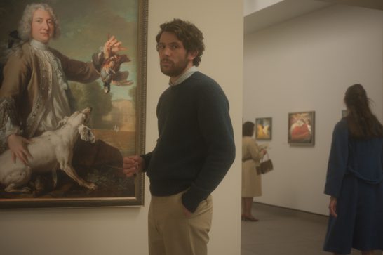 The focal point of the image is a large 18th-century portrait painting showing a well-dressed gentleman in period attire (powdered wig, ornate coat) holding what appear to be dead game birds - likely a hunting trophy portrait common among aristocracy of that era. The painting demonstrates the baroque or rococo style typical of European portraiture from that period. In the museum space, a man in contemporary casual clothing (dark sweater, light pants) stands looking at the painting, creating an interesting juxtaposition between past and present. In the background, another visitor can be seen viewing other artworks on the gallery walls.