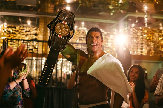A muscle bound man celebrates with a championship belt held high in what appears to be a festive, energetic atmosphere. He's wearing a white tank top and has a towel draped over his shoulder, surrounded by cheering people with golden confetti or lights creating a celebratory ambiance in the background. The championship belt appears to be a boxing or combat sports title, and the scene captures the joy and triumph of winning, possibly at a post-fight celebration or victory party.
