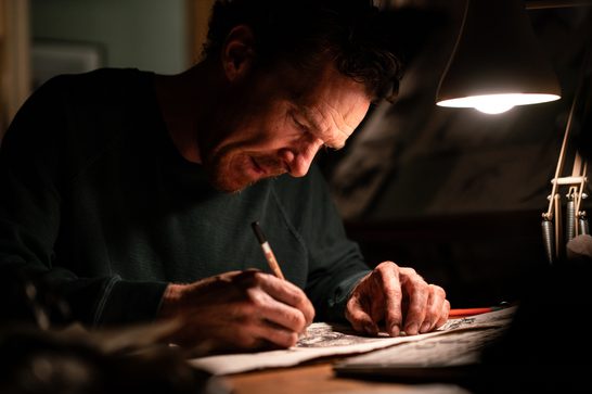 A bearded man in his late forties (Benedict Cumberbatch) crouches over a desk, concentrating as he scribbling away with a pencil on some paper. A desk lamp lights up his face.