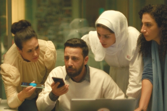 Four professionals are gathered around a laptop. A man in a white sweater is seated at the center, holding a phone while working on the laptop. Three women are standing around him, leaning in - one wearing a beige blouse on the left, one in a white hijab in the center-back, and one in blue on the right.