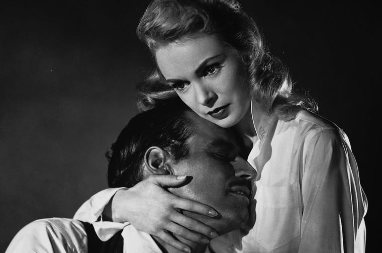 Black and white image of a younger woman with light hair holding an older man to her chest. The man has his eyes closed and the woman looks off to the left of camera.