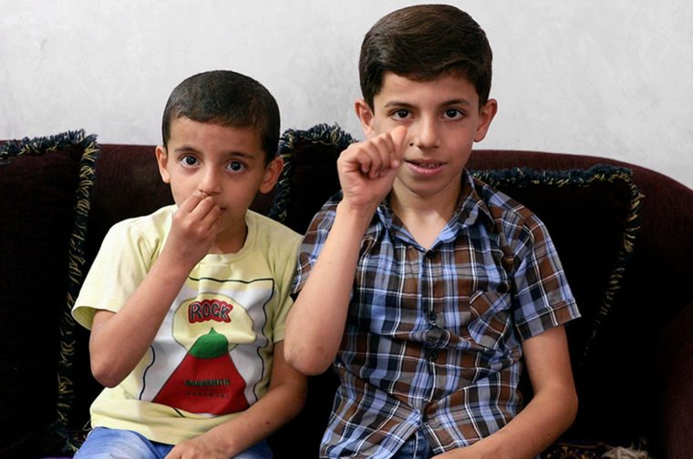 A photograph of two Palestinian young boys sitting together on a couch. The older boy on the right is wearing a plaid shirt and appears to be using sign language towards the camera, while the younger boy on the left in a yellow t-shirt with "ROCK" printed on it has his hand near his mouth in a shy pose. They're seated on what looks like a dark velvet couch with decorative fringe trim against a plain white wall.
