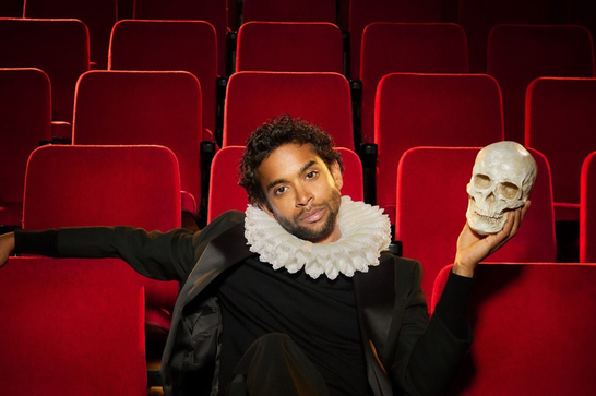 A South Asian man in his thirties sits in an audiotorium with plush red seats. He is wearing a white ruff, a black outfit and red socks, and his feet are crossed over the back of the chair in front of him. He looks directly into the camera. In his left hand he holds up a human skull.