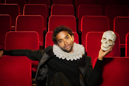 A South Asian man in his thirties sits in an audiotorium with plush red seats. He is wearing a white ruff, a black outfit and red socks, and his feet are crossed over the back of the chair in front of him. He looks directly into the camera. In his left hand he holds up a human skull.