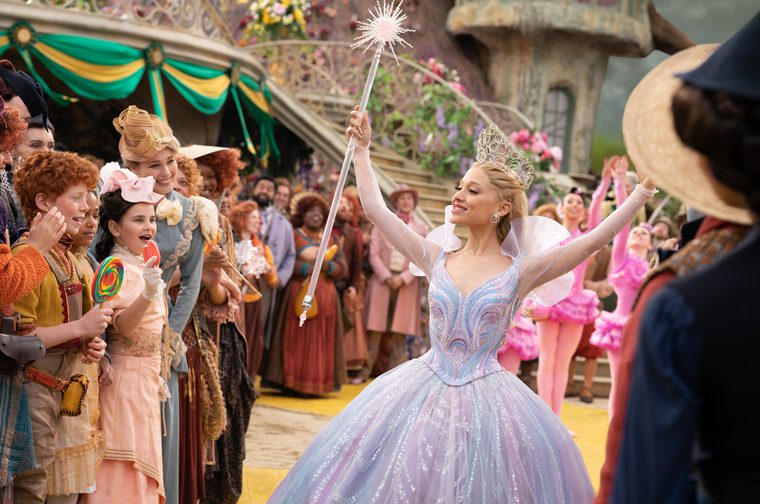 A fairy princess (Ariana Grande) holding a large magic wand with a star at the end holds her arms aloft, smiling at the people gathered around her. She is wearing a sparkly dress and has long braided blonde hair.