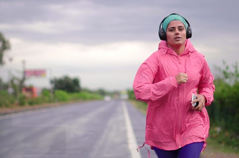 A South Asian woman in her late twenties jogs along a road, wearing a pink anorak, headphones and a blue head covering.