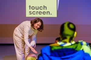 Woman with brunette bob and earing white top and trousers. She is leaning forward with her hand out. On the right is the back of a boy's head. The screen in the background has a yellow box on it with the word 'touch screen'.