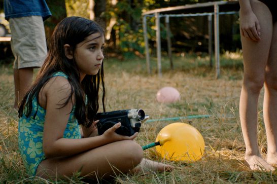 This still frame shows a young girl with long dark hair sitting cross-legged in dry summer grass, wearing a teal and green floral swimsuit. She holds a small camcorder in both hands and gazes off to the side with a serious, focused expression. Beside her on the grass is a large yellow water balloon attached to a hose, with a pink balloon visible further back. Partially visible around her are other children's legs, and in the blurred background stands a trampoline frame and trees lit by warm, golden light suggesting late afternoon or early evening.