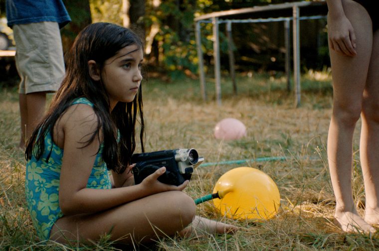 This still frame shows a young girl with long dark hair sitting cross-legged in dry summer grass, wearing a teal and green floral swimsuit. She holds a small camcorder in both hands and gazes off to the side with a serious, focused expression. Beside her on the grass is a large yellow water balloon attached to a hose, with a pink balloon visible further back. Partially visible around her are other children's legs, and in the blurred background stands a trampoline frame and trees lit by warm, golden light suggesting late afternoon or early evening.