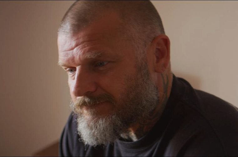 The image shows a middle-aged man with a shaved/closely cropped grey head, a full grey-white beard, and a visible tattoo on the side of his neck. He's wearing a dark shirt and appears to be in a contemplative or serious mood, looking slightly downward.