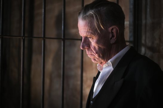 Close-up profile of an older man in a dark jacket and open-collared white shirt, standing in front of a barred window. Warm light falls across his deeply lined face while the rest of the scene stays in shadow. His gray hair is slicked back with a few loose strands falling forward, and his gaze is lowered and intense.