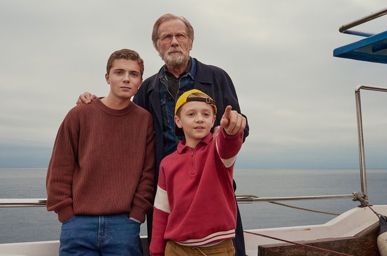 An older man with a grey beard and glasses stands on the deck of a boat with his arm around a teenage boy in a red sweater and jeans, while a young child in a red and white top and yellow cap points excitedly into the distance. A calm grey sea and overcast sky stretch behind them.