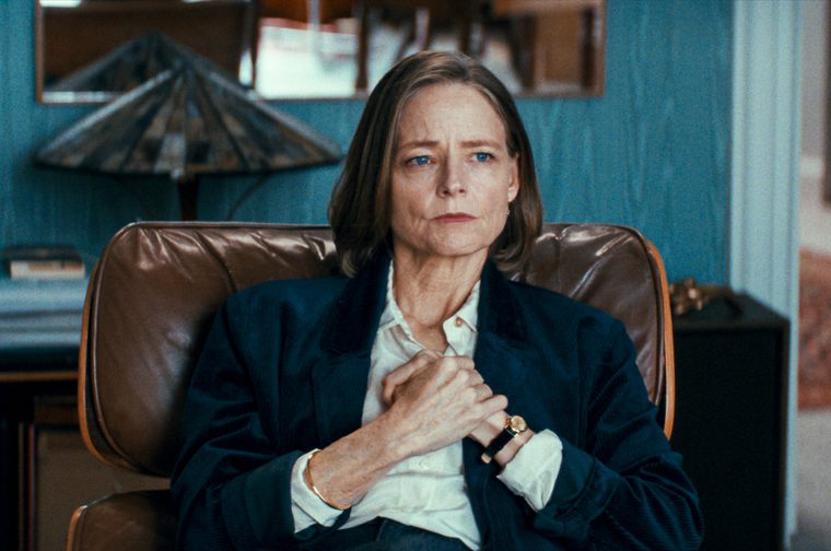 A middle-aged woman with shoulder-length brown hair and blue eyes sits in a brown leather armchair, hands clasped together at her chest. She wears a navy blazer over a white button-up shirt and a gold watch. Her expression is calm and composed. Behind her is a bookshelf filled with binders and books, a Tiffany-style lamp, and blue-painted walls. A patterned rug is partially visible to the right.