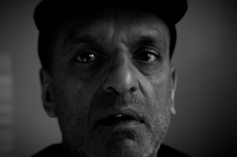 The image is a striking black-and-white close-up portrait of a middle-aged Asian man. He's wearing a dark baseball cap and has a short beard. The lighting is dramatic and moody, with deep shadows emphasizing the texture of his face. His expression is intense and somewhat unsettled — eyes wide, mouth slightly open.