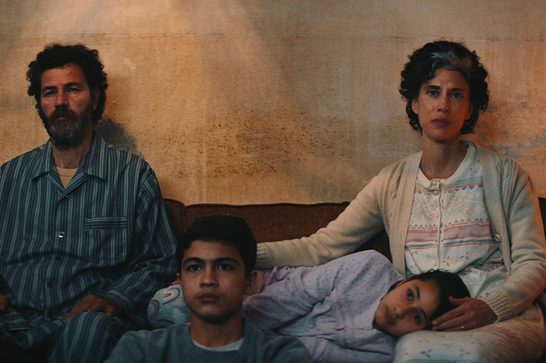 The image shows a sombre family scene with two adults and two children sitting on a bed against a weathered wall, capturing the intimate and difficult circumstances portrayed in the film. One of the children is resting their head on their mother's lap. The warm, muted color palette and naturalistic setting are characteristic of the film's visual style.