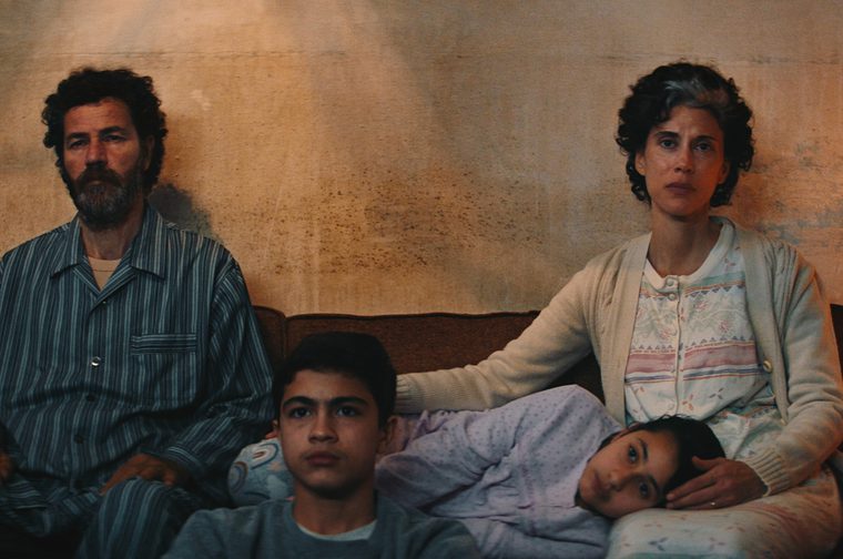 The image shows a sombre family scene with two adults and two children sitting on a bed against a weathered wall, capturing the intimate and difficult circumstances portrayed in the film. One of the children is resting their head on their mother's lap. The warm, muted color palette and naturalistic setting are characteristic of the film's visual style.