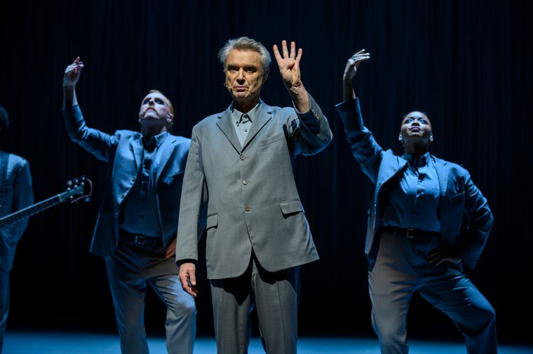An older male performer (David Byrne) in a light grey suit stands center stage, raising one hand with fingers spread, wearing a headset microphone. Behind him, two backing performers in darker grey outfits strike dynamic poses with arms raised, against a dark draped backdrop. A guitar neck is visible at the far left edge of the frame.