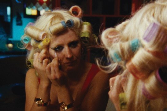 A woman with blonde hair set in colorful curlers leans on her hand while applying eyeliner, reflected in a mirror in warm, dim lighting.