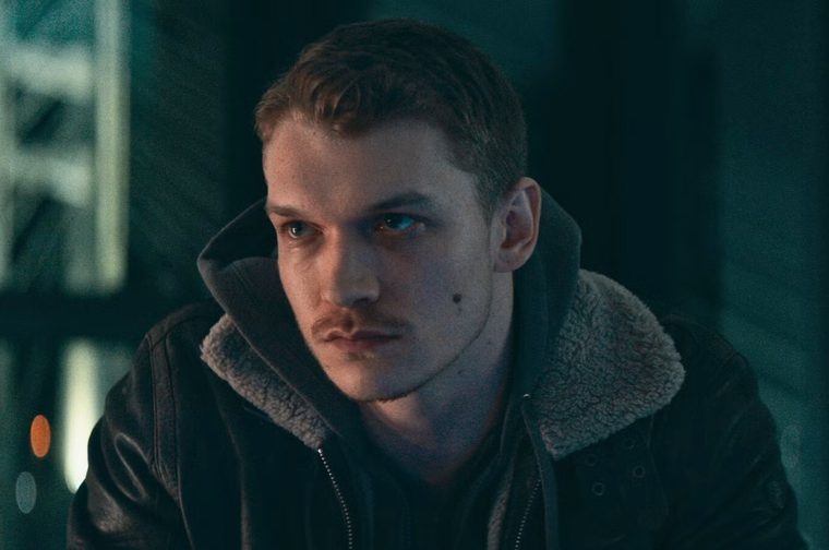A young man with short, wavy reddish-brown hair and light stubble sits looking off to the side with a pensive expression. He wears a dark leather jacket with a shearling-lined collar over a black hoodie. The background is dimly lit in cool teal and dark tones, with blurred structural elements and a few soft orange and white lights visible, suggesting an industrial or urban night setting.