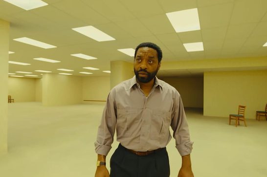 A Black man in his late forties wearing a light purple button-down shirt and dark trousers stands in a vast, empty room with yellowish walls, fluorescent panel lighting in the ceiling, and sparse wooden chairs in the background. He looks alert and slightly uneasy, as if disoriented in the liminal, maze-like space.