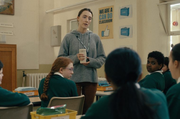 A young woman in a grey hoodie with a staff lanyard stands at the front of a primary school classroom, holding a coffee cup, addressing a group of students in green uniforms. Behind her, a wall displays a 'We Are a Can Do Classroom' poster and handwritten sentence strips about a cow and spilled milk.