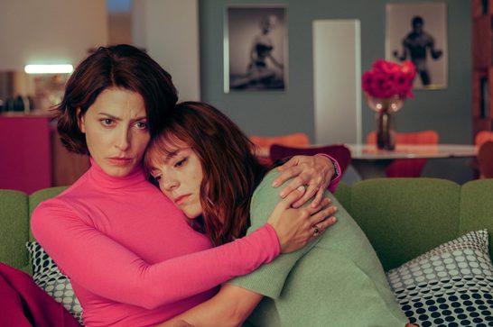 Two women embrace on a green sofa in a stylish, colorful living room. One, with short dark hair and a pink turtleneck, holds a worried expression while comforting the other, who has long auburn hair and wears a pink cardigan over a sage green top, resting her head on the first woman's shoulder with a troubled look. The room features black-and-white photographs on gray walls, a vase of pink roses, and warm ambient lighting.