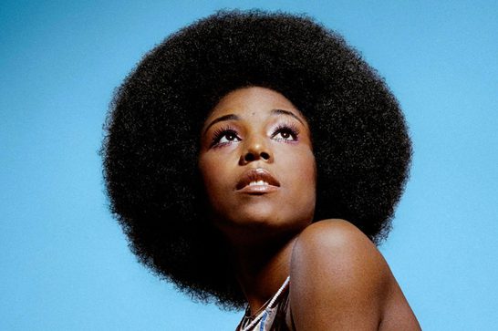 A striking studio portrait of a woman with a large, voluminous natural afro, photographed against a solid sky-blue background. She gazes upward, chin lifted, with defined eye makeup and glossy lips. Her bare shoulder and a beaded necklace are visible in the lower right of the frame.