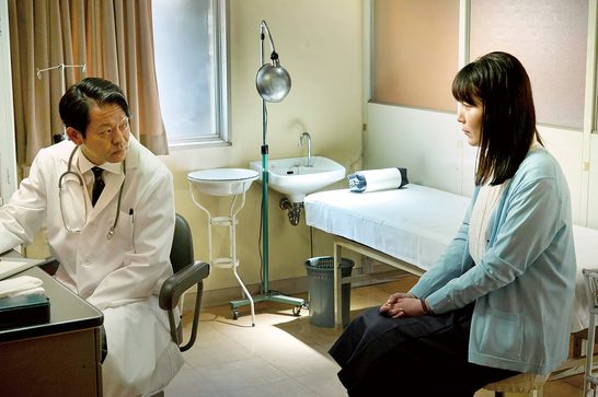 A Japanese doctor in a white lab coat with a stethoscope sits across from a female Japanese patient in a medical examination room. Between them are a sink on a stand and an adjustable lamp. An examination table with a white paper cover is visible in the background.