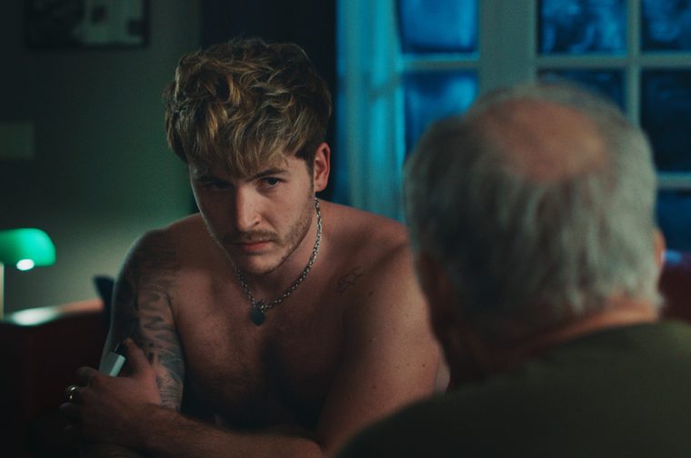 A shirtless young man with tousled blond hair, a mustache, tattooed arm, and a silver chain necklace sits leaning forward, gazing intently at an older man seated across from him with his back to the camera. The scene is lit moodily with a green desk lamp and blue light glowing through French doors in the background.