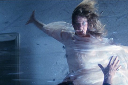 A possessed woman with wild, wind-blown hair appears mid-air in a state of violent motion, arms flung wide, her face contorted in a terrified scream with one eye rolled back. She's dressed in a pale, flowing garment that ripples as if caught in a powerful gust, set against a dim, textured blue-gray wall with an open doorway to the left.