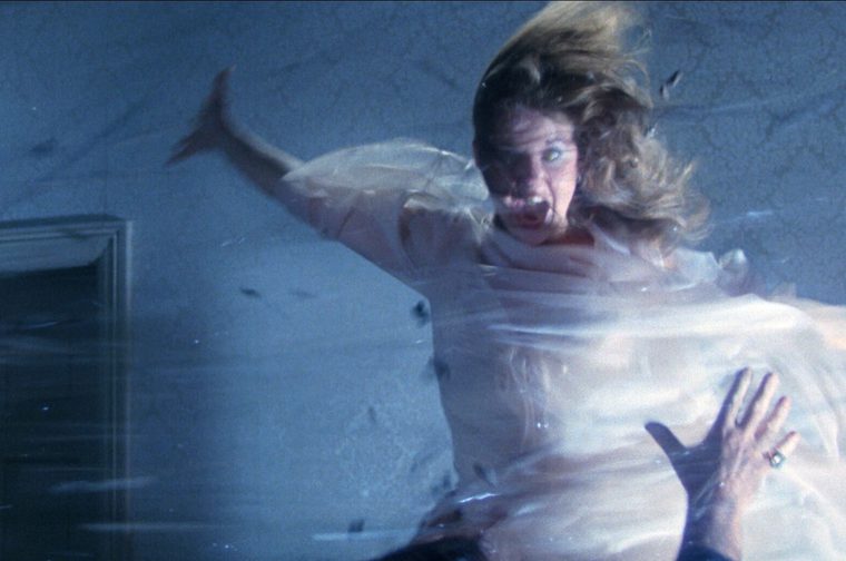 A possessed woman with wild, wind-blown hair appears mid-air in a state of violent motion, arms flung wide, her face contorted in a terrified scream with one eye rolled back. She's dressed in a pale, flowing garment that ripples as if caught in a powerful gust, set against a dim, textured blue-gray wall with an open doorway to the left.