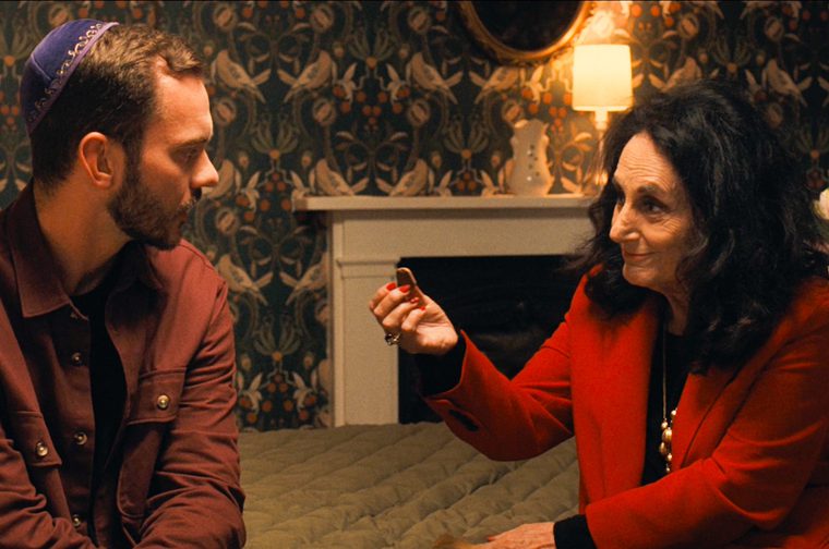 In a cosy bedroom with ornate bird-and-pomegranate wallpaper, a young bearded man in a purple embroidered kippah and a burgundy overshirt sits on a quilted bed, glancing sideways at an older woman beside him. She has voluminous dark hair, red nail polish and a bright red blazer, and holds a small biscuit-like piece of food between her fingers with a wry, affectionate smile. A lit lamp, a white fireplace, a vase of pink lilies and a brass bedpost frame the scene.