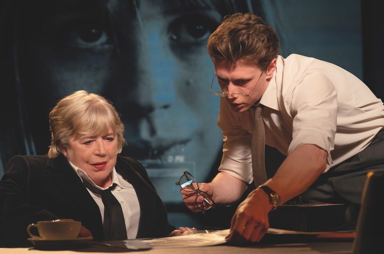 An older woman with short blonde hair (Marianna Faithfull), dressed in a black blazer and tie, sits at a desk examining papers. A younger man in a white dress shirt and loosened tie leans over the desk beside her, holding glasses in one hand and pointing at documents with the other. A large, dramatic close-up of a face is projected on the backdrop behind them.