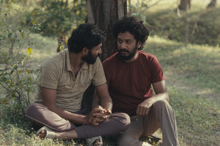 Two bearded South Asian men sit together on the ground leaning against a tree trunk in a lush green outdoor setting. The man on the left wears a beige striped polo shirt and cross-legged with sandals, while the man on the right wears a dark red t-shirt with his legs extended. They are facing each other in conversation, their hands clasped together between them.