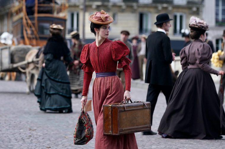 A young woman in late 19th-century dress stands on a cobblestone street, carrying a leather suitcase and a tapestry bag. She wears a red polka-dot blouse with puffed sleeves, a long dusty-rose skirt, a blue sash belt, and a small straw hat. She gazes to her left amid a bustling crowd of period-dressed pedestrians, with horse-drawn carriages and a stone building visible in the background.