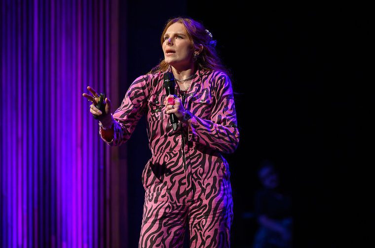 We can see a lady in her twenties on stage holding a microphone, wearing a striking pink and black zebra-print jumpsuit. They have long auburn hair with hair clips, layered necklaces, and rings, and are gesturing expressively with one hand. The stage is lit with purple and blue lighting against a dark background.
