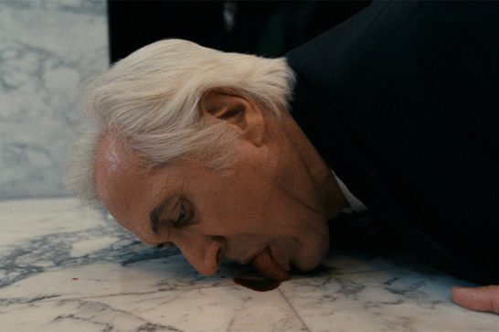 The image shows an older man with white hair, dressed in a dark suit, crouched down with his face pressed to a white marble floor, licking a small pool of dark red blood.