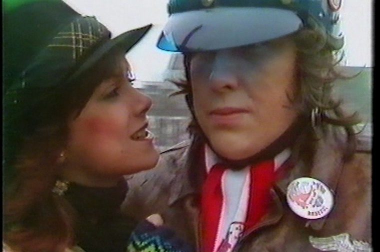 We see a younger man and woman (both white) in what appears to be a vintage photograph, likely from the 1970s or 1980s based on the clothing and video quality. One person is wearing a plaid/tartan hat and colorful clothing, while the other is wearing a peaked cap/visor pulled down low over their face, a red scarf or neckerchief, a brown jacket, and a round badge/pin on their lapel. The image has the characteristic grainy, slightly washed-out look of VHS or early video footage.