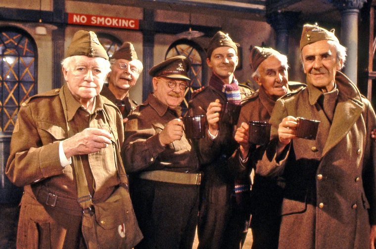Six members of the Home Guard platoon from classic sitcom Dad's Army stand in a row raising mugs in a toast, dressed in Home Guard uniforms and forage caps, in front of a shop with a 'No Smoking' sign.