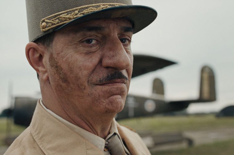 Close-up of an older moustached man in a pale khaki uniform and tie, wearing a tall kepi decorated with gold leaf embroidery. He glances at the camera with a firm, unsmiling look. Behind him, a dark olive military bomber with roundel markings sits on a grassy airfield under an overcast sky.