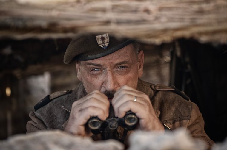 Close-up of an older mustached man in a military beret with a badge and a khaki uniform with shoulder epaulettes. He looks intently through a pair of binoculars, framed by the blurred timber and sandbag-like edges of a trench or bunker. A wedding ring is visible on his hand.