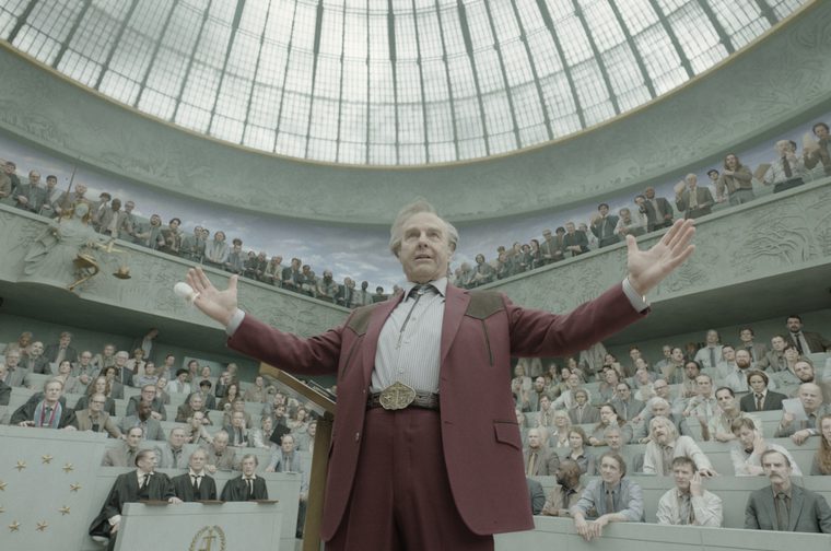 An older man in a maroon Western-style suit with a bolo tie and large ornate belt buckle stands at a podium with arms outstretched, addressing a packed, tiered courtroom or assembly hall. The chamber has a grand domed glass ceiling and stone walls carved with reliefs, with a statue of blindfolded Justice holding scales visible on the upper level. Rows of onlookers, judges, and officials fill the surrounding balconies and gallery seats, all facing him.