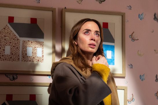 A woman with reddish-brown hair and tearful, red-rimmed eyes looks up with an anguished, frightened expression, clutching the sleeves of a yellow sweater and brown coat close to her chest. She stands in a room with pink butterfly-patterned wallpaper, framed folk-art-style house illustrations with red roofs and patterned facades hanging on the wall beside her.